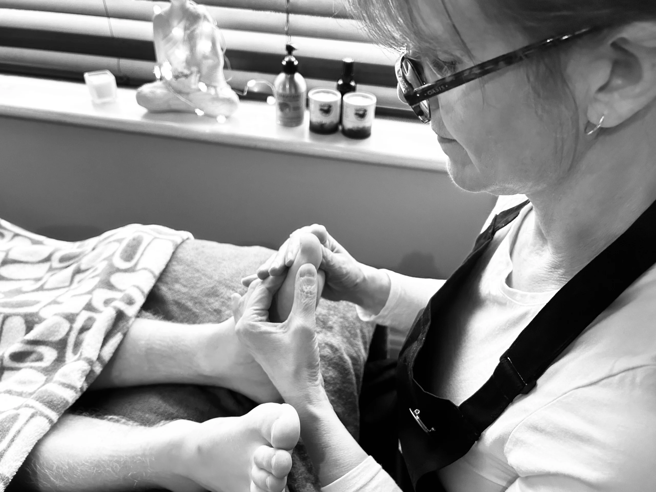 Francesca, in a dark apron and glasses, working on a client's foot; candles and oil bottles sit on the windowsill behind.